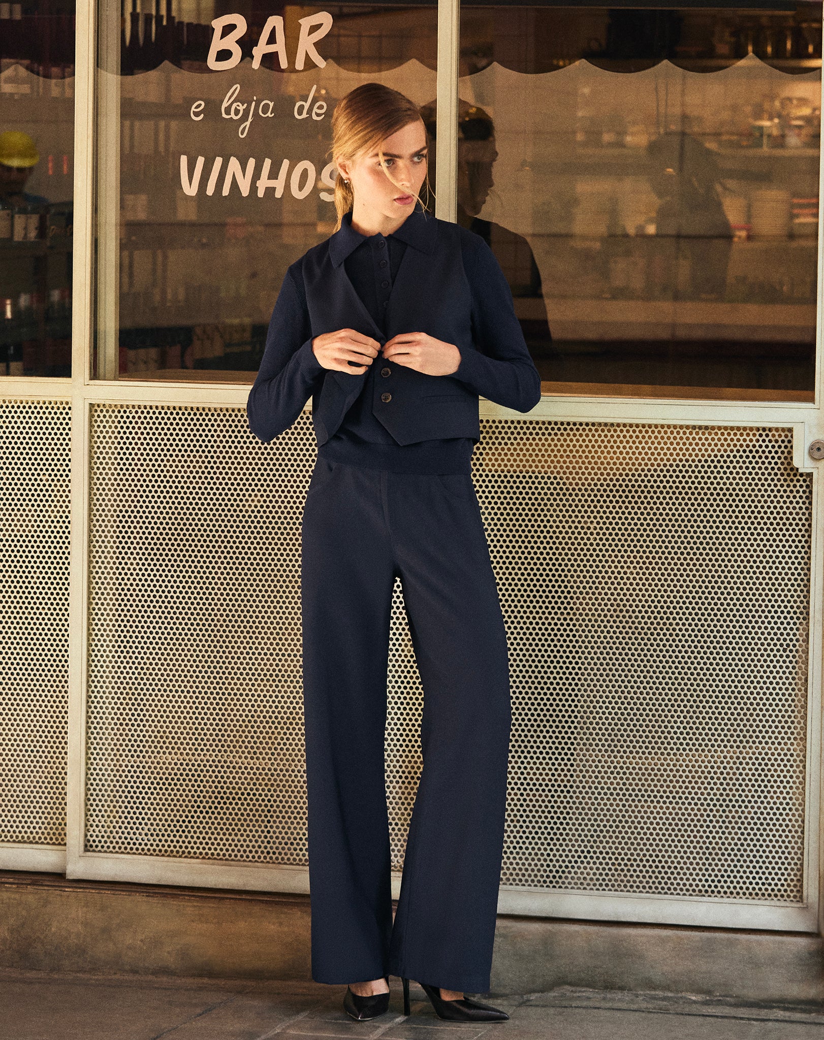 Modelo usando um suéter gola polo de tricô azul marinho com calça bege - estilo corpo inteiro, mangas longas com caimento regular, foto em ambiente urbano.