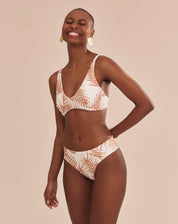 Modelo vestindo top de biquíni estampado com palmeiras em tons camelo sobre fundo areia, vista frontal.