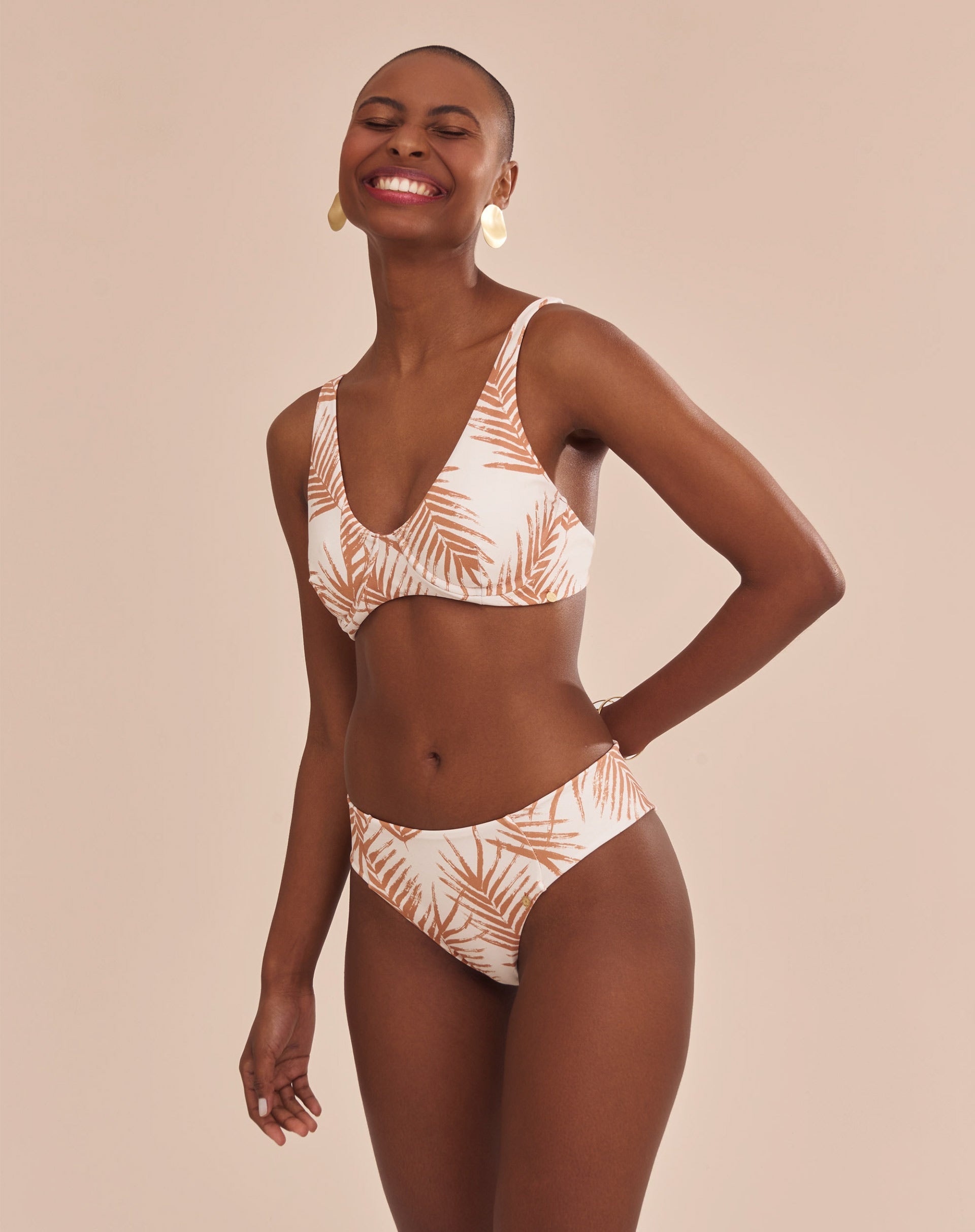 Modelo vestindo top de biquíni estampado com palmeiras em tons camelo sobre fundo areia, vista frontal.