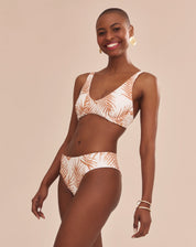 Modelo vestindo top de biquíni estampado com palmeiras em tons camelo sobre fundo areia, vista lateral.