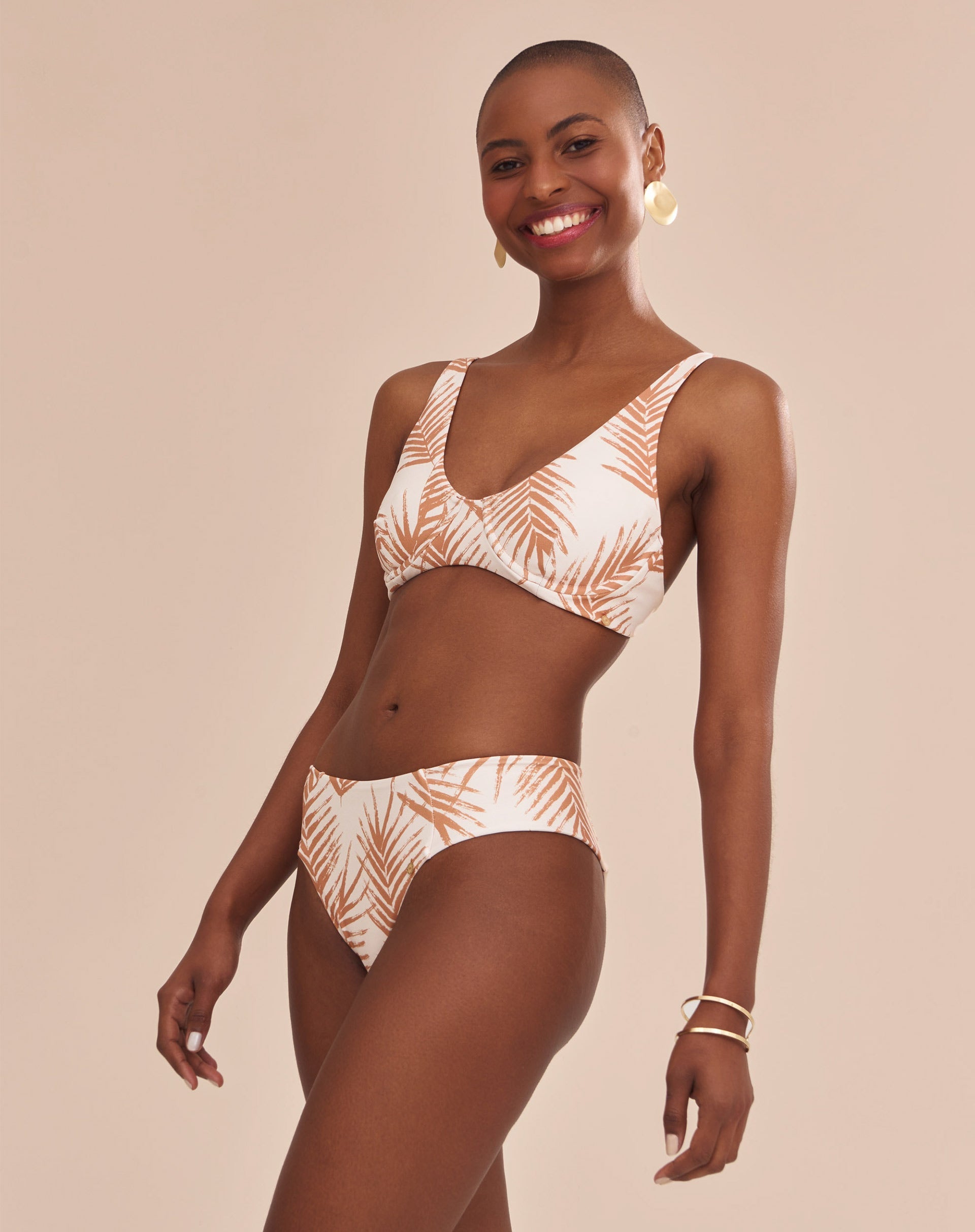 Modelo vestindo top de biquíni estampado com palmeiras em tons camelo sobre fundo areia, vista lateral.