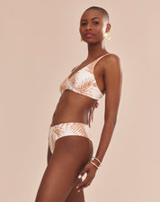 Modelo vestindo top de biquíni estampado com palmeiras em tons camelo sobre fundo areia, vista traseira.