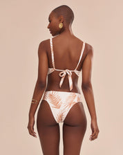 Modelo vestindo top de biquíni estampado com palmeiras em tons camelo sobre fundo areia, vista lateral traseira.