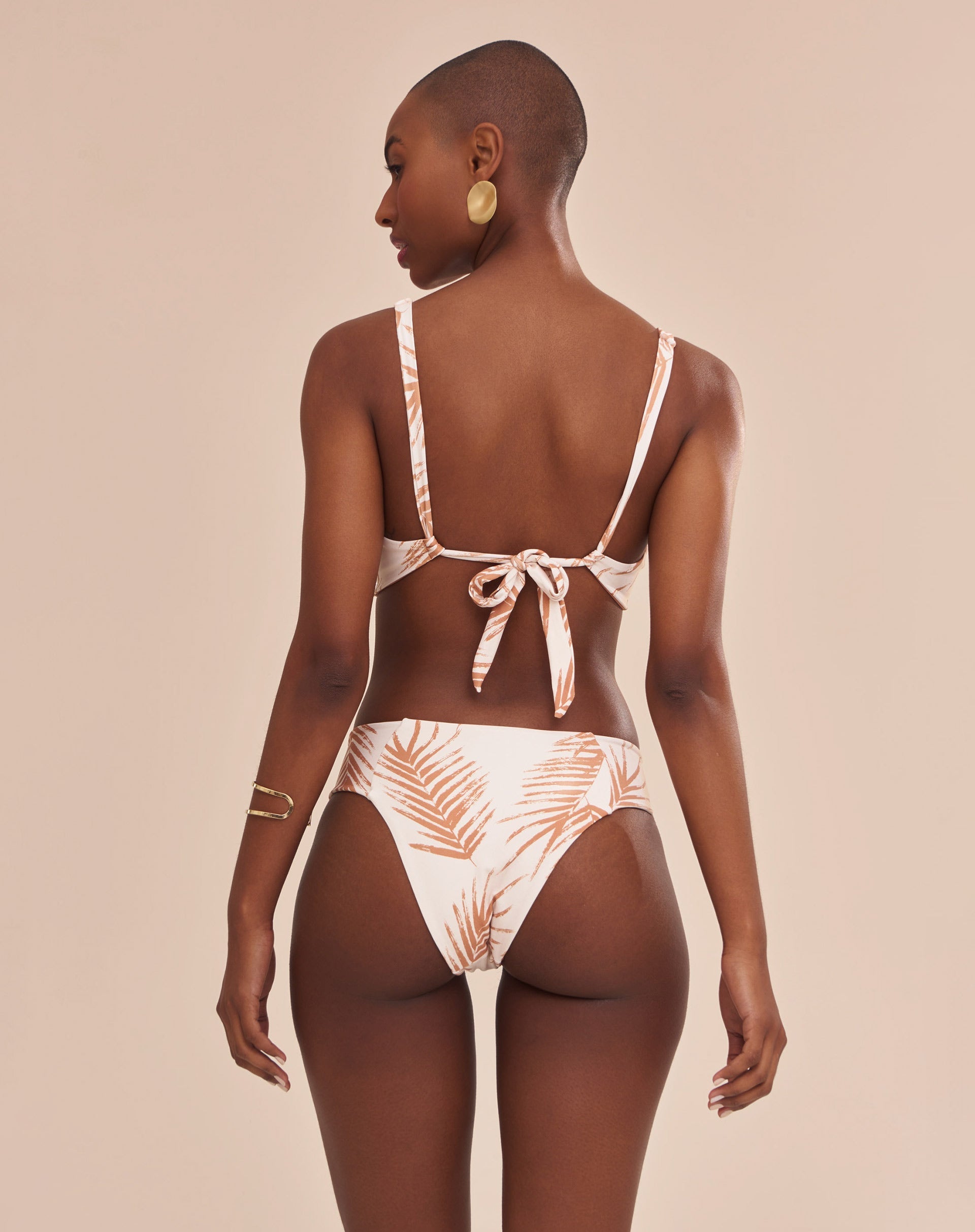 Modelo vestindo top de biquíni estampado com palmeiras em tons camelo sobre fundo areia, vista lateral traseira.