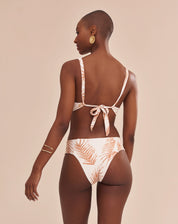 Modelo vestindo top de biquíni estampado com palmeiras em tons camelo sobre fundo areia, vista frontal.