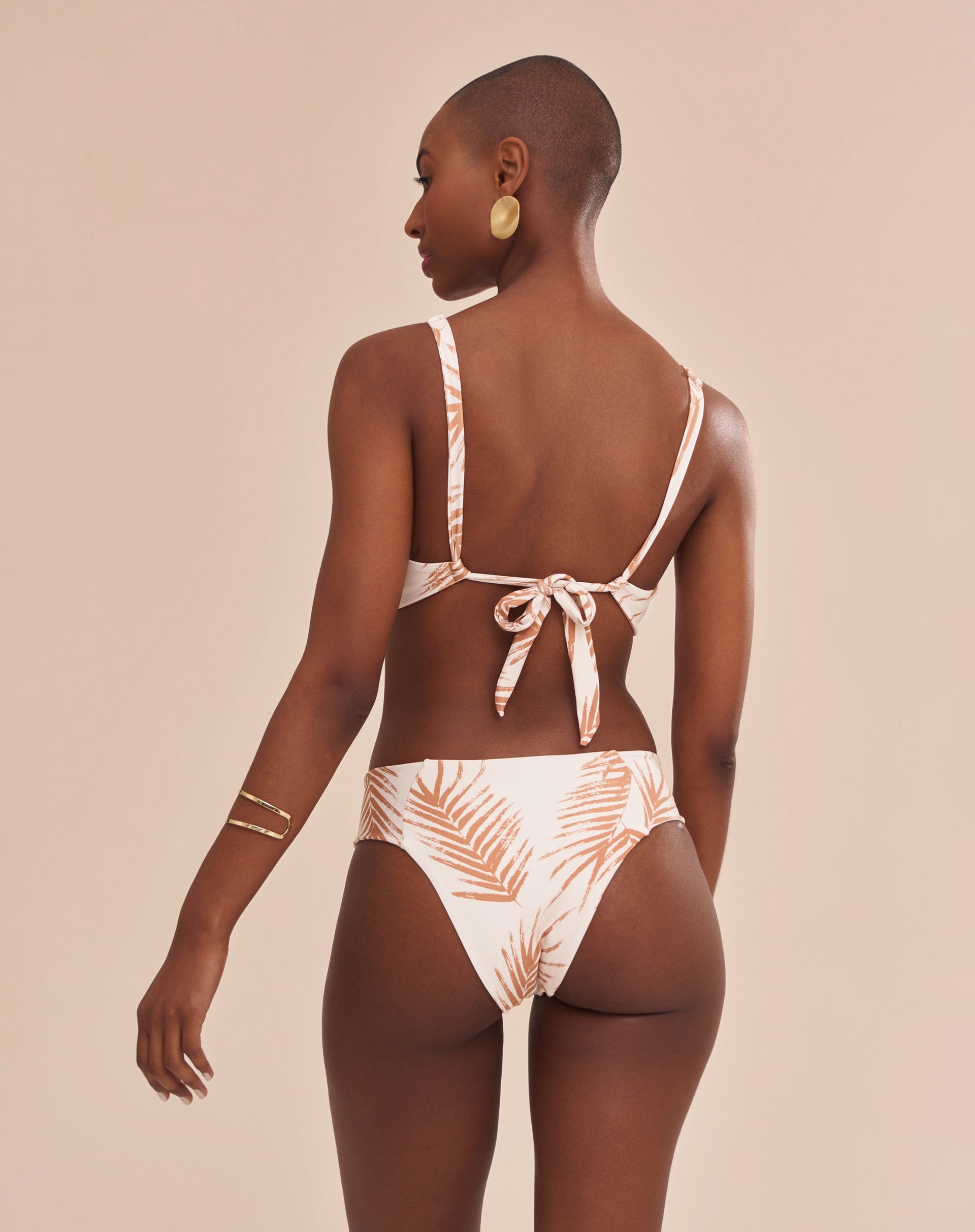Modelo vestindo top de biquíni estampado com palmeiras em tons camelo sobre fundo areia, vista frontal.