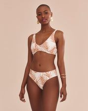 Modelo vestindo top de biquíni estampado com palmeiras em tons camelo sobre fundo areia, vista lateral.