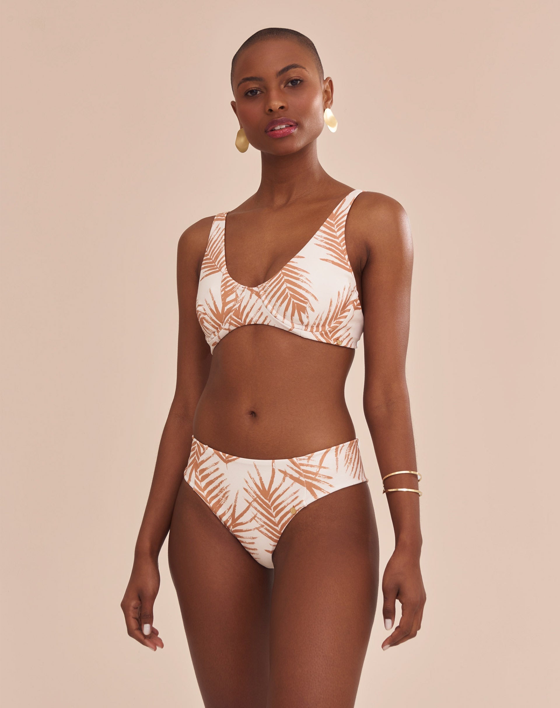 Modelo vestindo top de biquíni estampado com palmeiras em tons camelo sobre fundo areia, vista lateral.