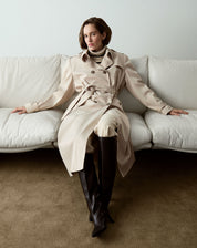 Modelo sentado em sofá vestindo trench coat bege com botas de couro.
