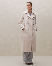Modelo em pé vestindo trench coat bege com calças cinza e sapatos pretos.