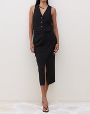 Modelo vestindo saia midi preta com fenda frontal e colete preto, em pose frontal.