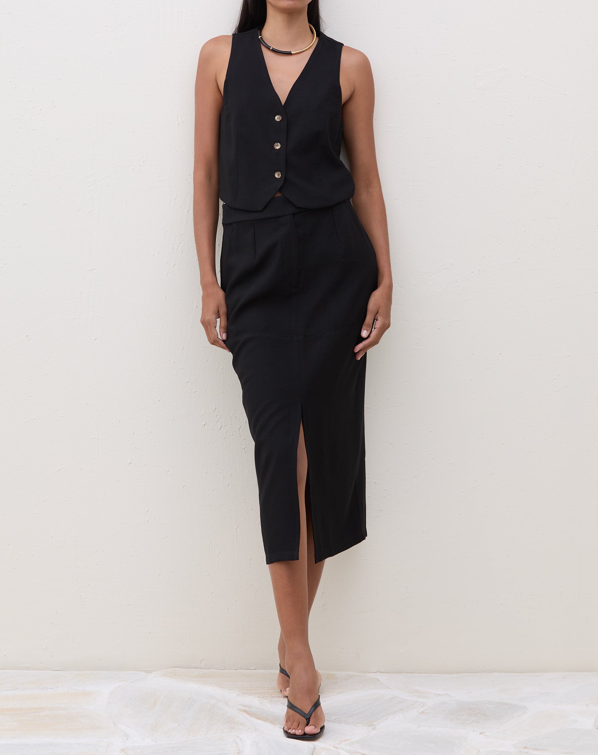 Modelo vestindo saia midi preta com fenda frontal e colete preto, em pose frontal.