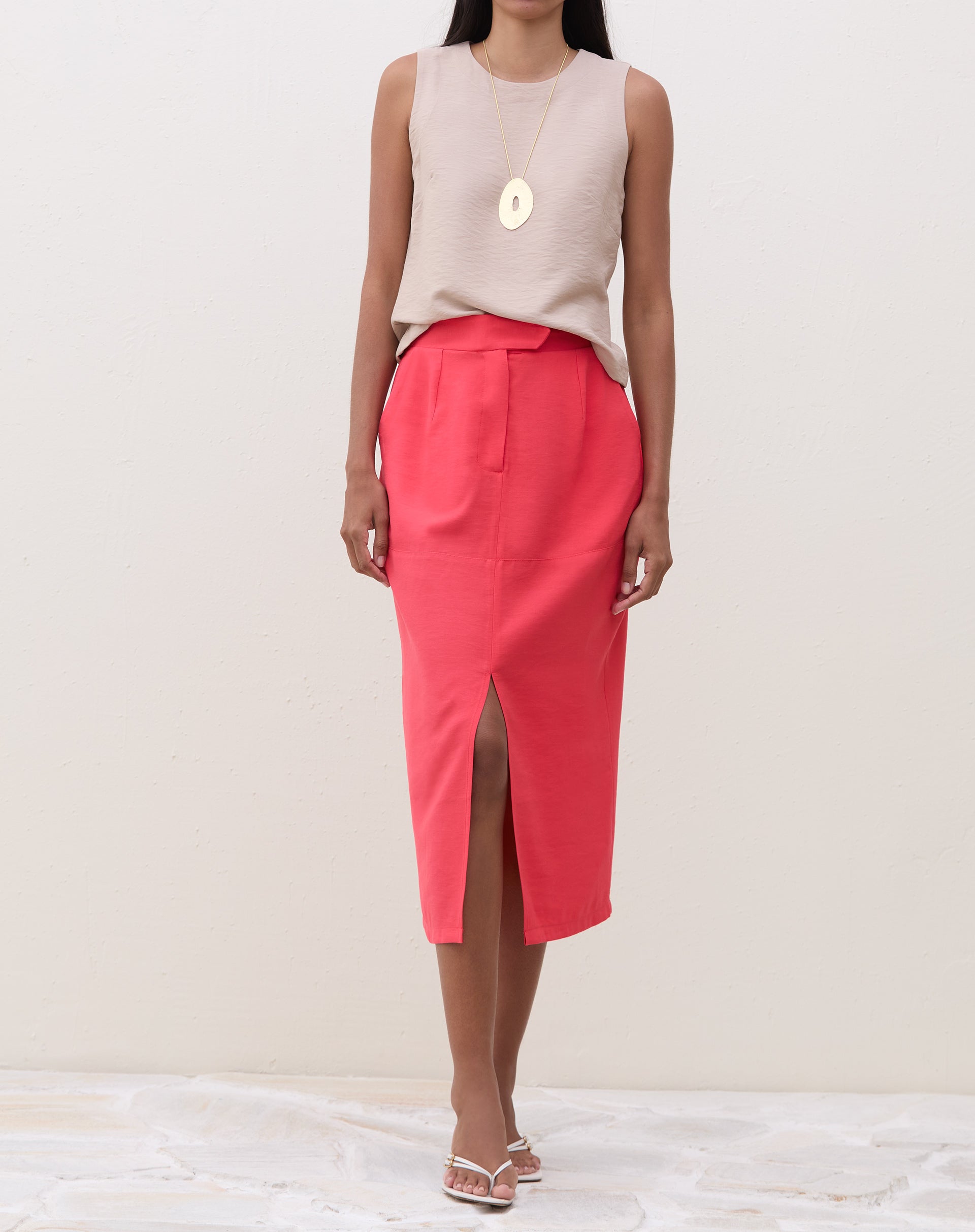 Modelo vestindo saia midi coral com fenda frontal, combinada com blusa neutra e sandálias.