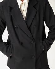 Close-up de corpo mostrando abotoamento duplo do blazer oversized preto, lapela entalhada e bolsos embutidos com mão no bolso.