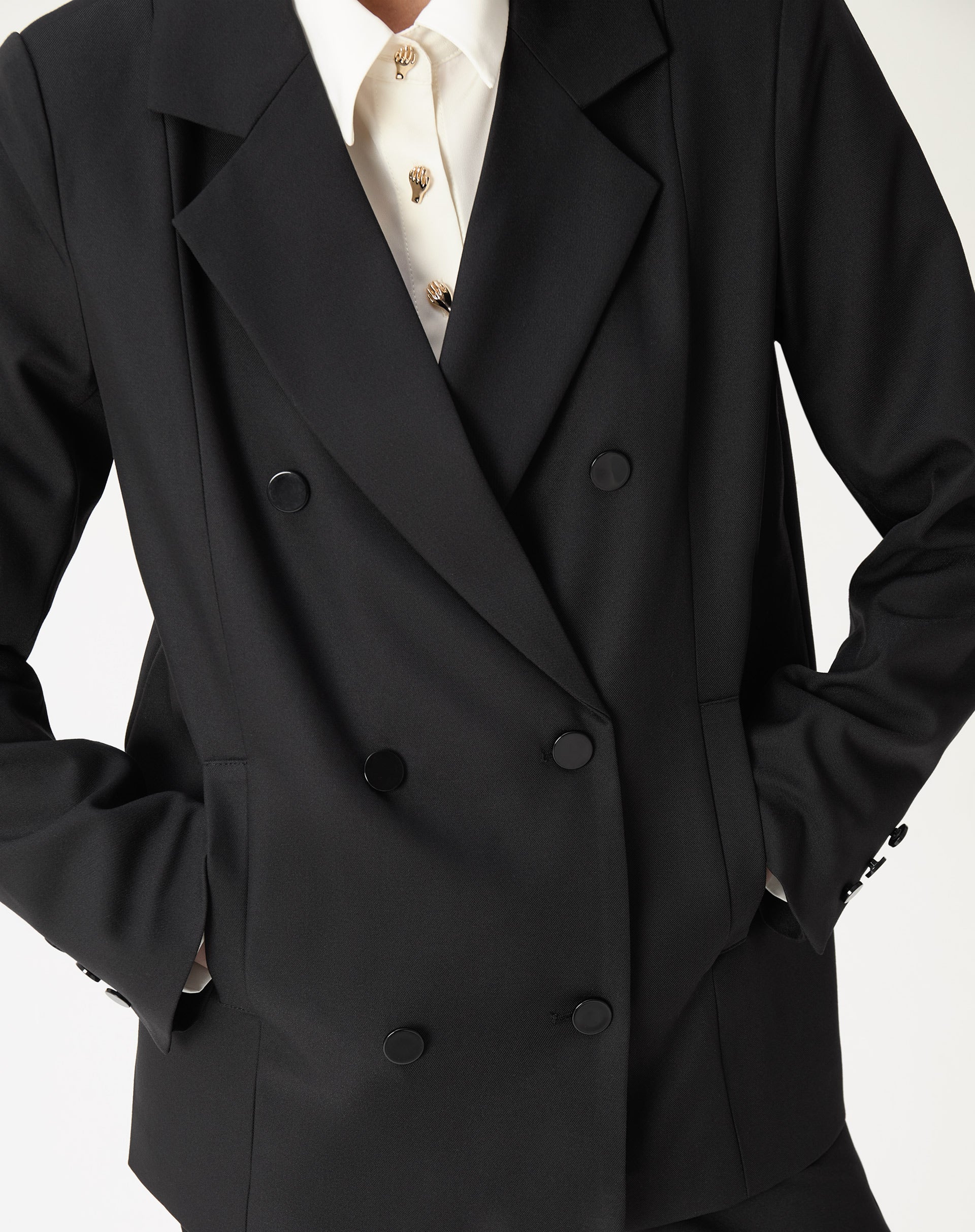 Close-up de corpo mostrando abotoamento duplo do blazer oversized preto, lapela entalhada e bolsos embutidos com mão no bolso.