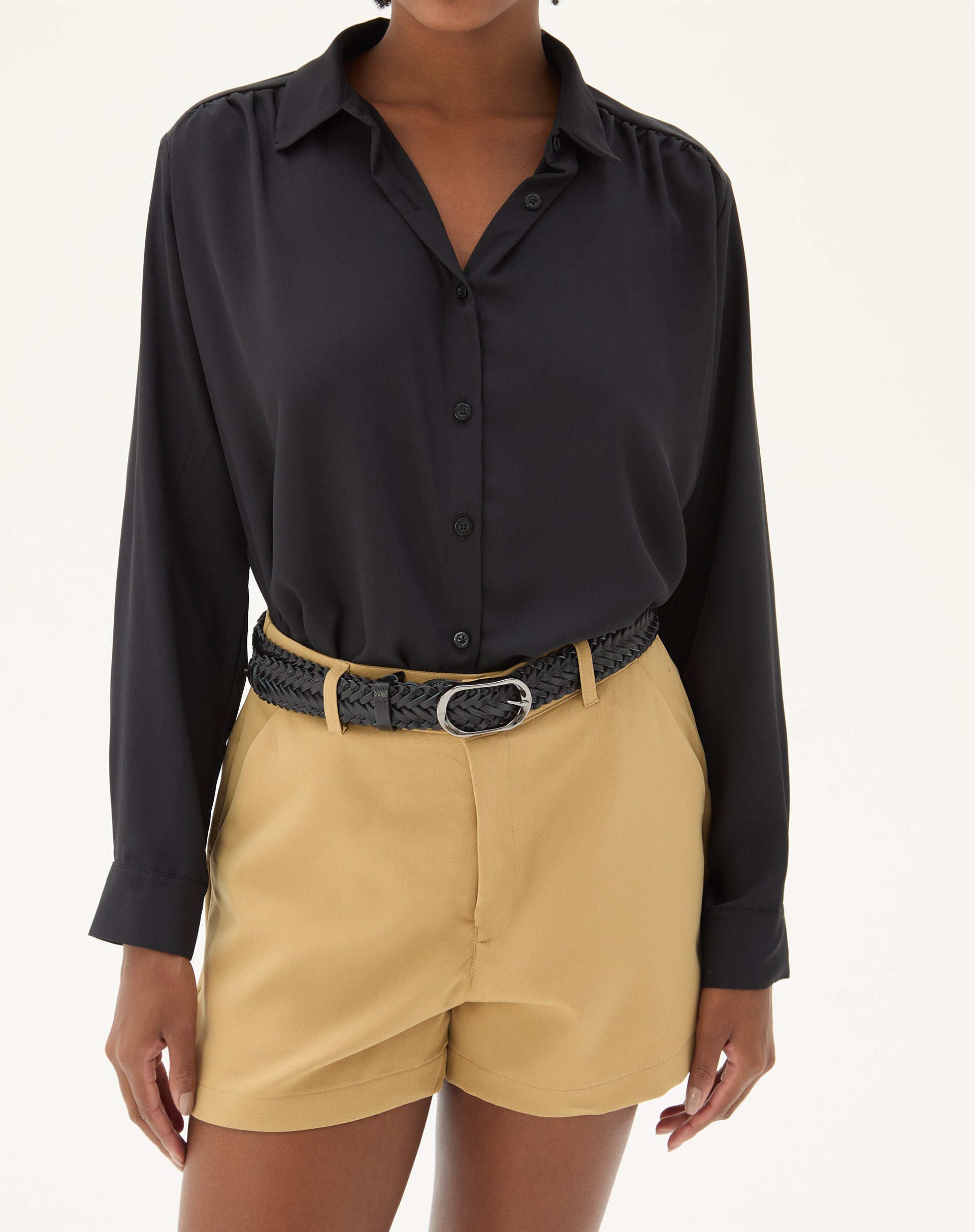 Modelo vestindo shorts curto reto de alfaiataria na cor areia com cinto preto e camisa preta.