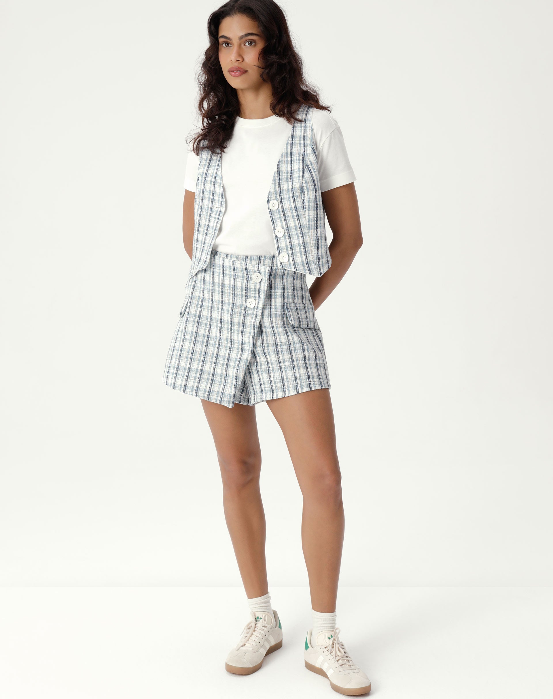 Modelo vestindo shorts frente transpassada de alfaiataria com estampa xadrez e tecido tweed, combinado com camiseta branca e tênis.