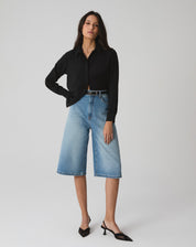 Modelo vestindo camisa preta de manga longa com barra reta, combinada com bermuda jeans e sapatos pretos.