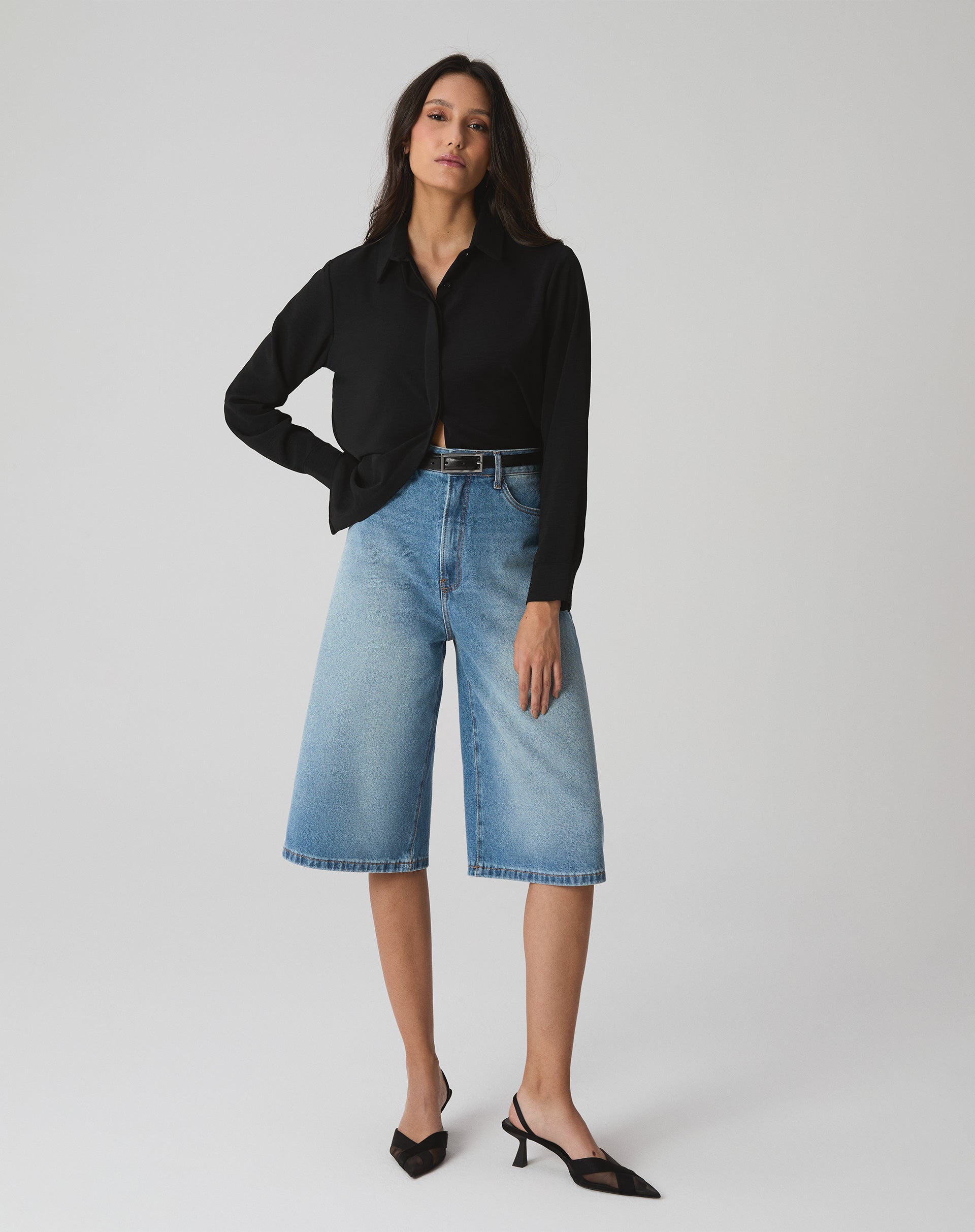 Modelo vestindo camisa preta de manga longa com barra reta, combinada com bermuda jeans e sapatos pretos.