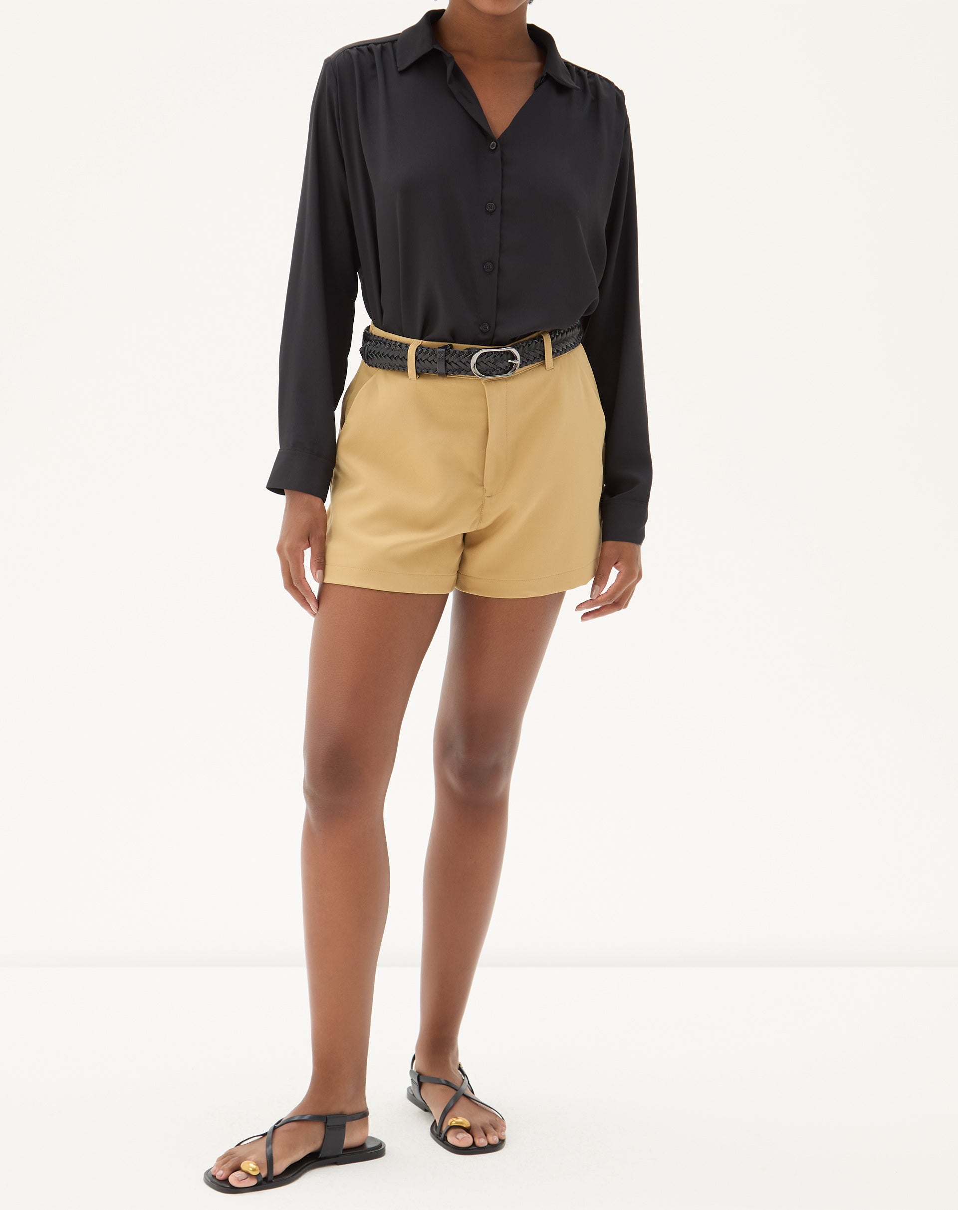 Modelo vestindo camisa preta de manga longa com barra reta, combinada com shorts bege e sandálias pretas.