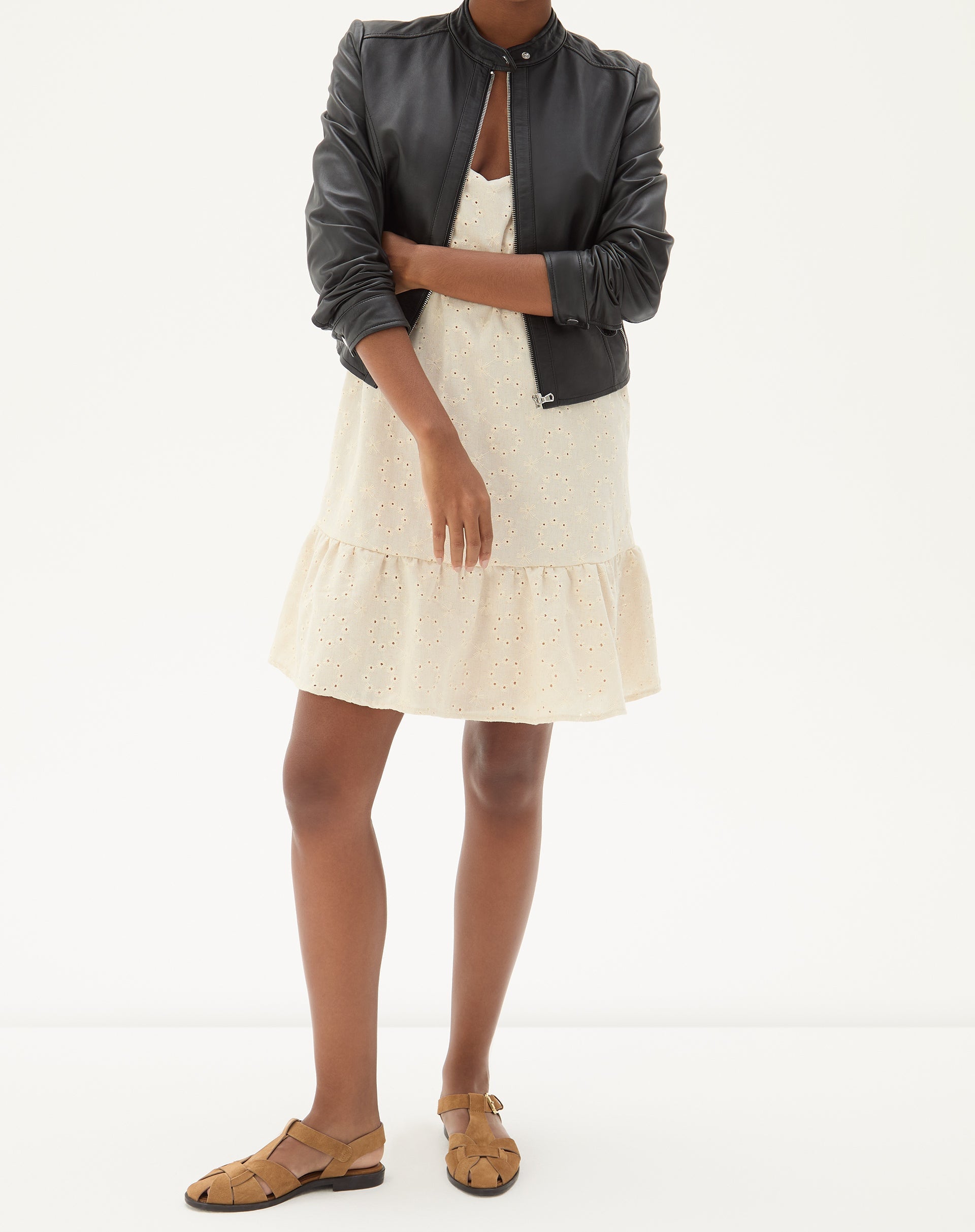 Modelo vestindo vestido curto bege com alças finas, combinado com jaqueta preta e sandálias marrons.