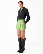 Modelo em pose lateral vestindo shorts verde com vincos e camisa preta, destacando o look elegante.