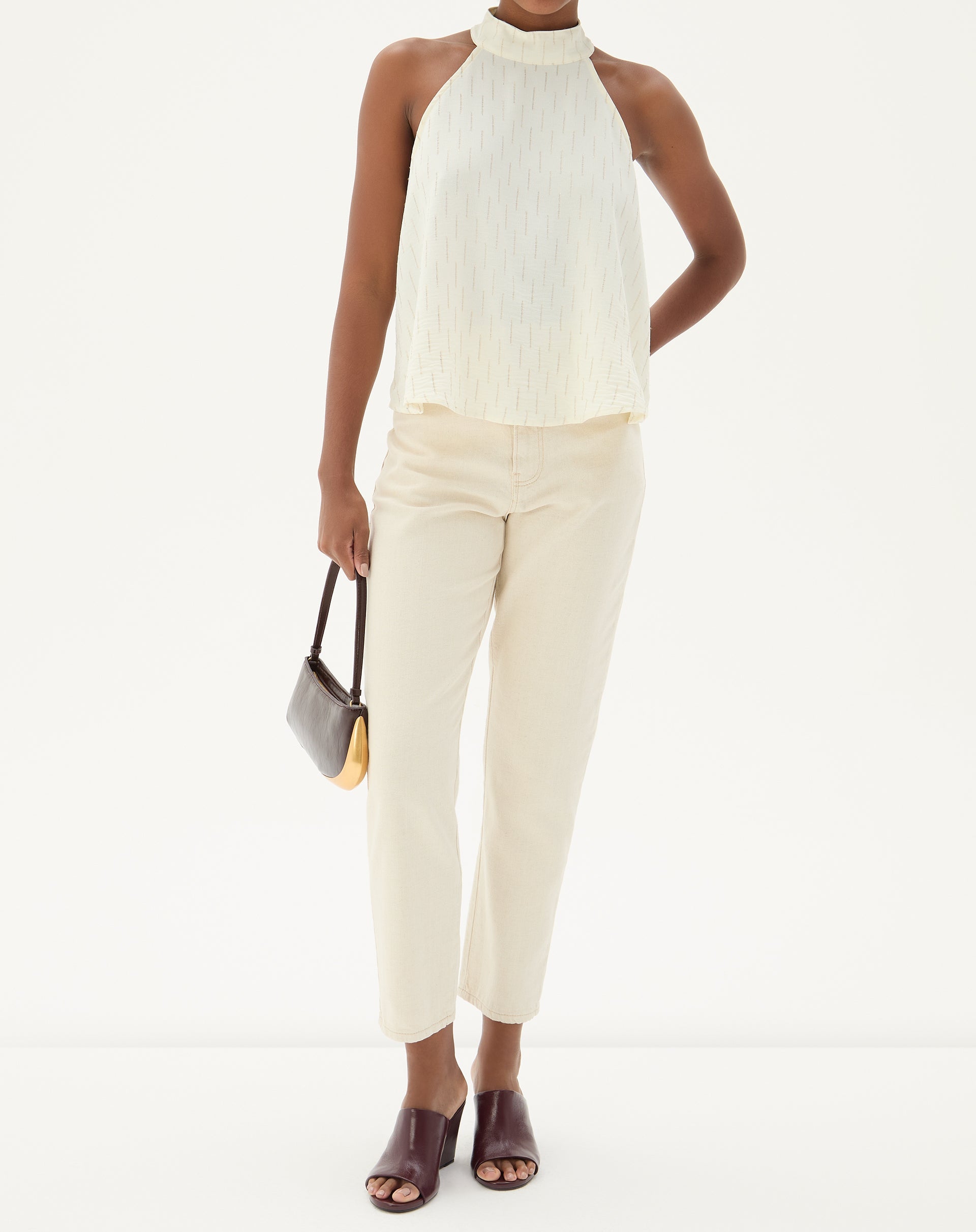 Modelo vestindo calça bege com blusa sem mangas e sandálias, segurando uma bolsa.