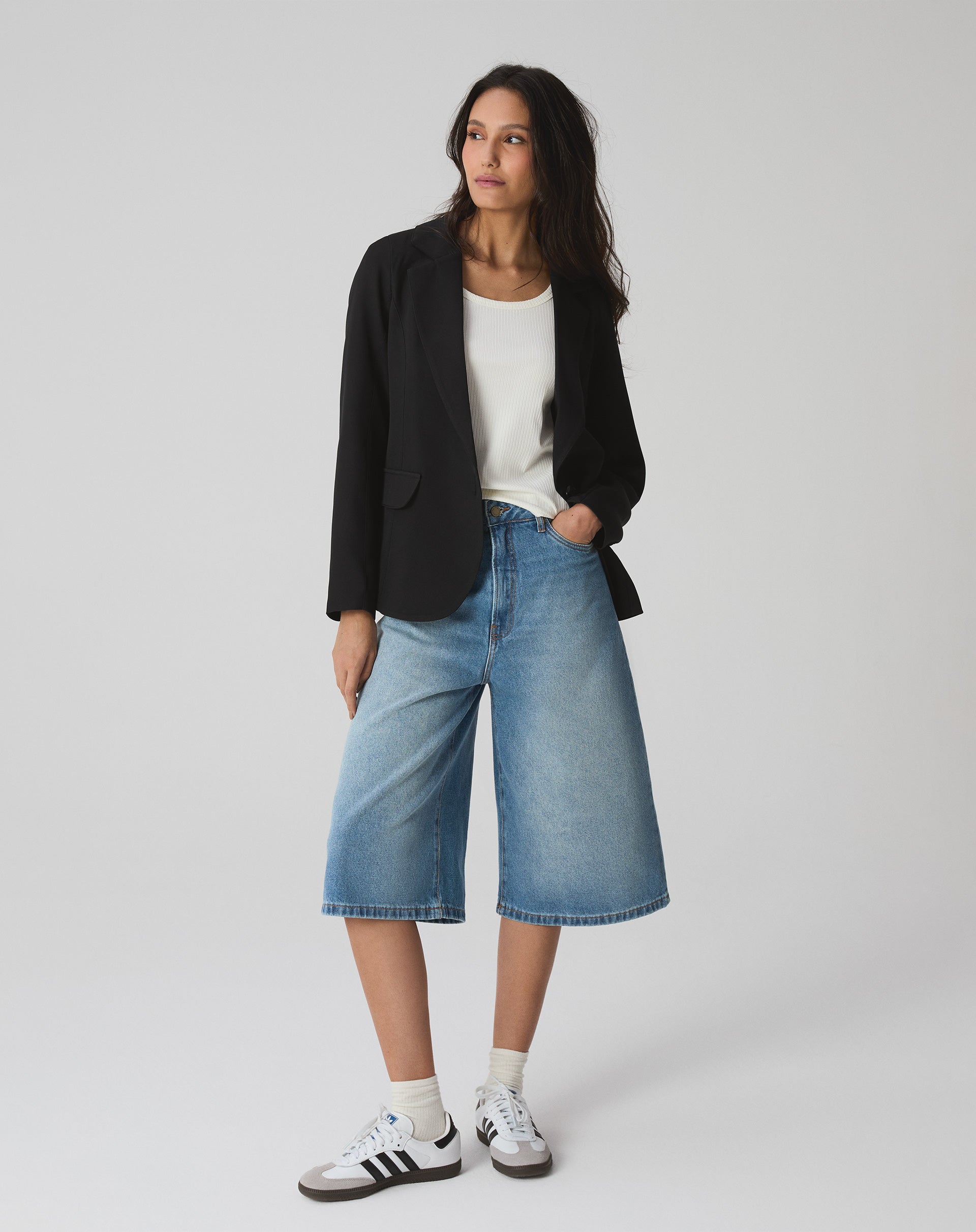 Modelo vestindo bermuda jeans alongada evasê com blazer preto e tênis branco.