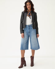 Modelo vestindo bermuda jeans alongada evasê com jaqueta preta e botas marrons.