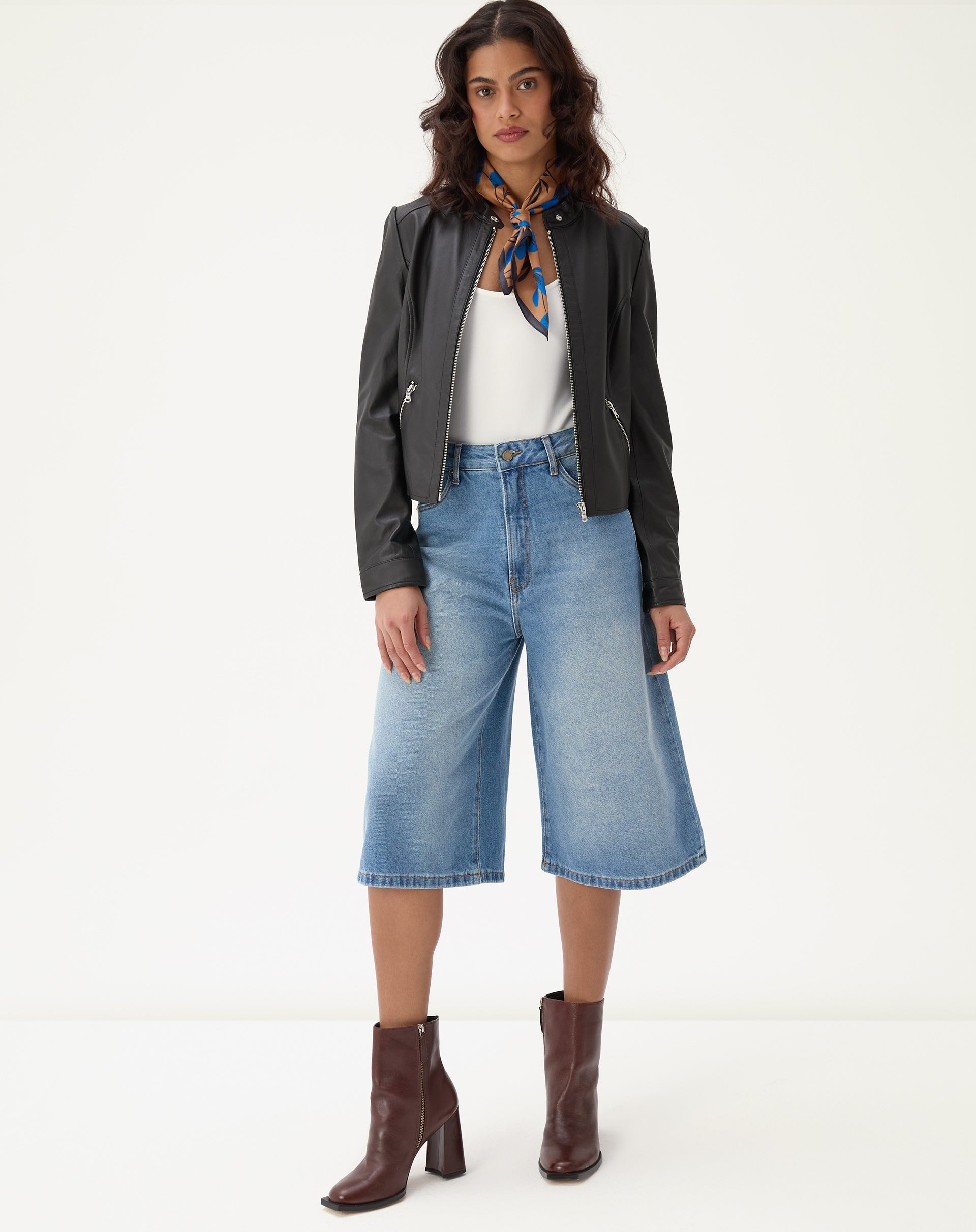 Modelo vestindo bermuda jeans alongada evasê com jaqueta preta e botas marrons.