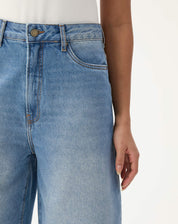 Close-up da frente da bermuda jeans alongada evasê mostrando detalhes do cós e botão.