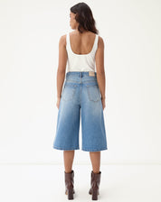 Vista traseira da bermuda jeans alongada evasê sendo usada por modelo com botas marrons.