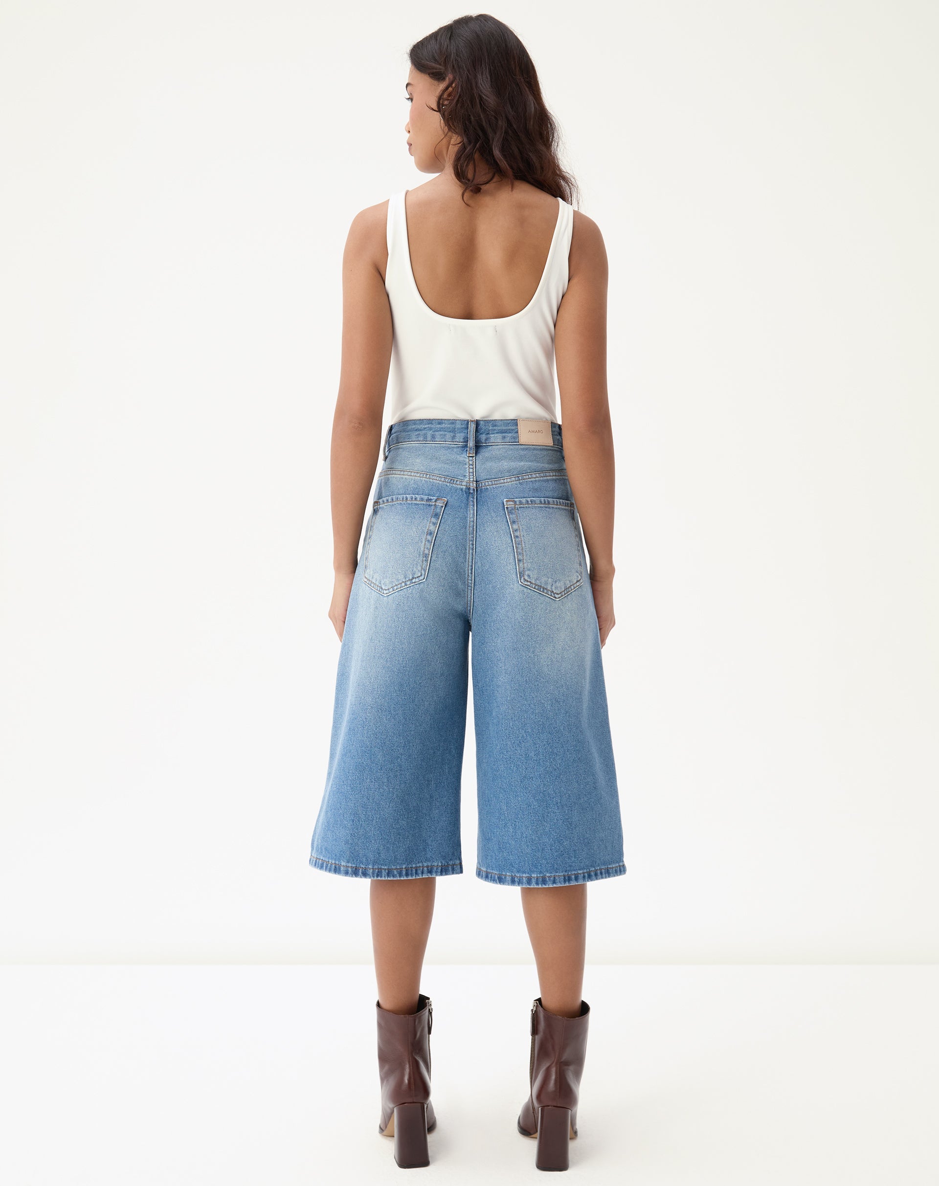 Vista traseira da bermuda jeans alongada evasê sendo usada por modelo com botas marrons.