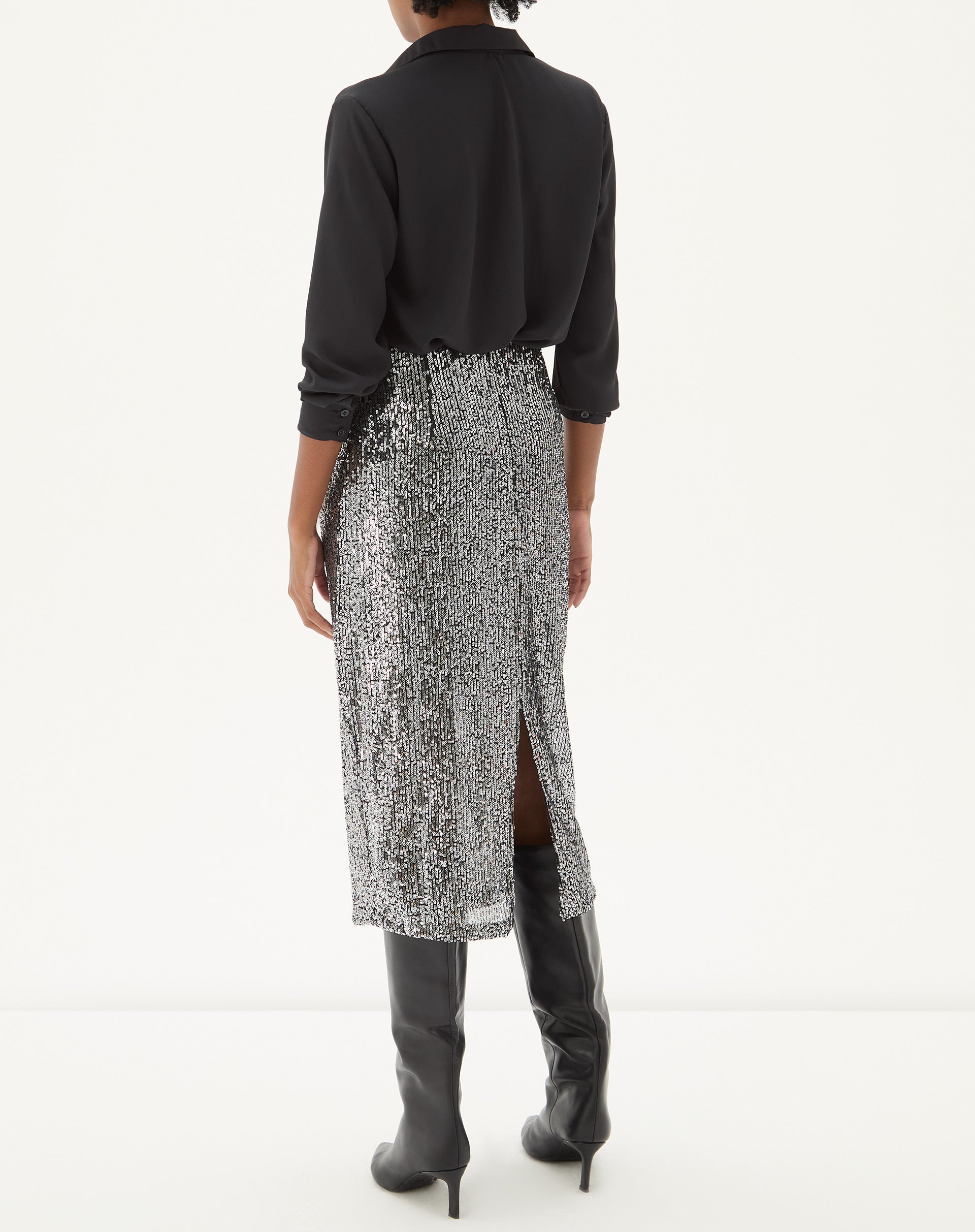 Modelo vestindo a Saia Midi Tubo de Paetê - Prata com camisa preta e botas pretas, vista traseira.