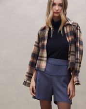 Modelo usando shorts azul com vivo contrastante, combinado com blusa preta e jaqueta xadrez.