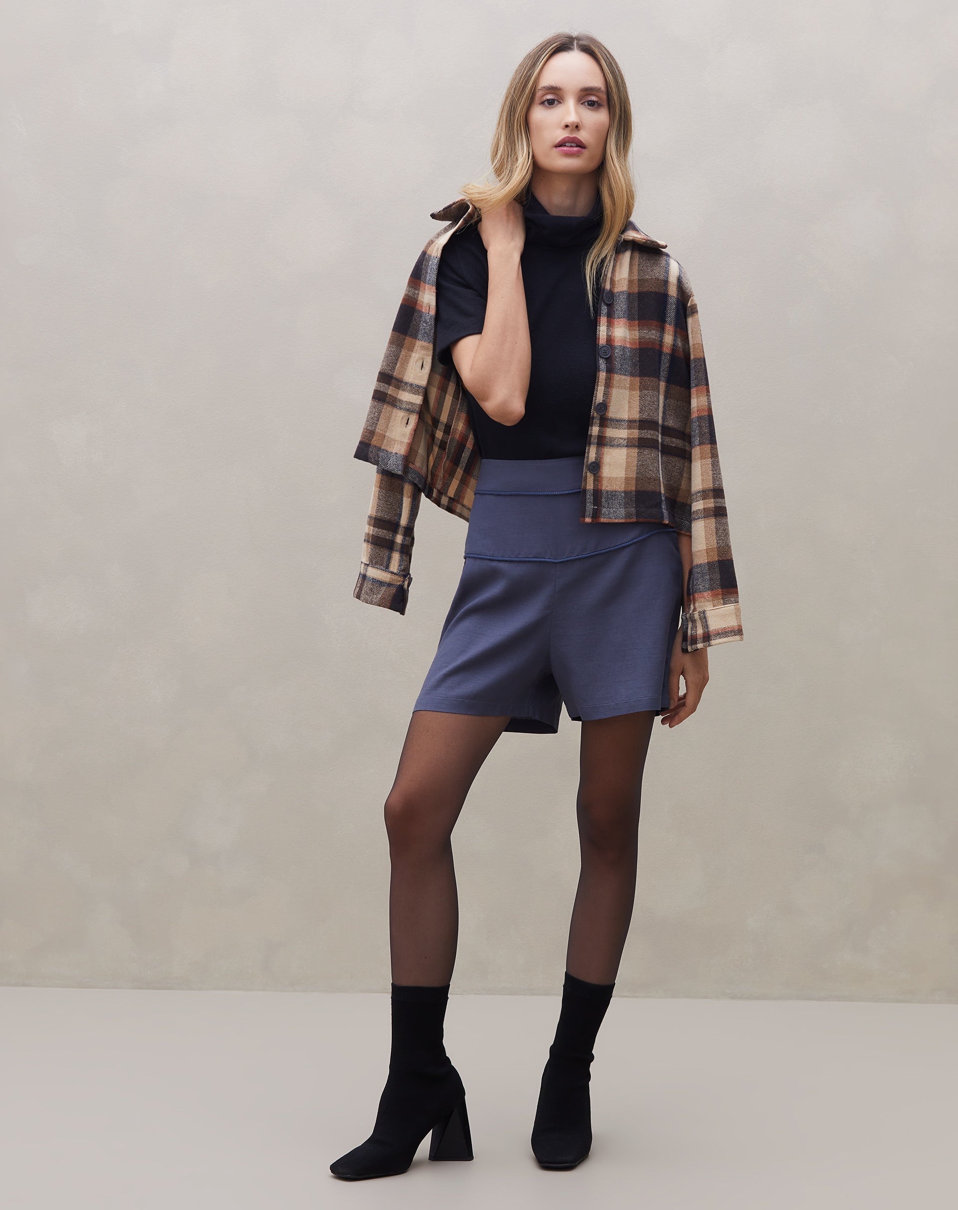 Modelo vestindo shorts azul com vivo contrastante, combinado com blusa preta e jaqueta xadrez.