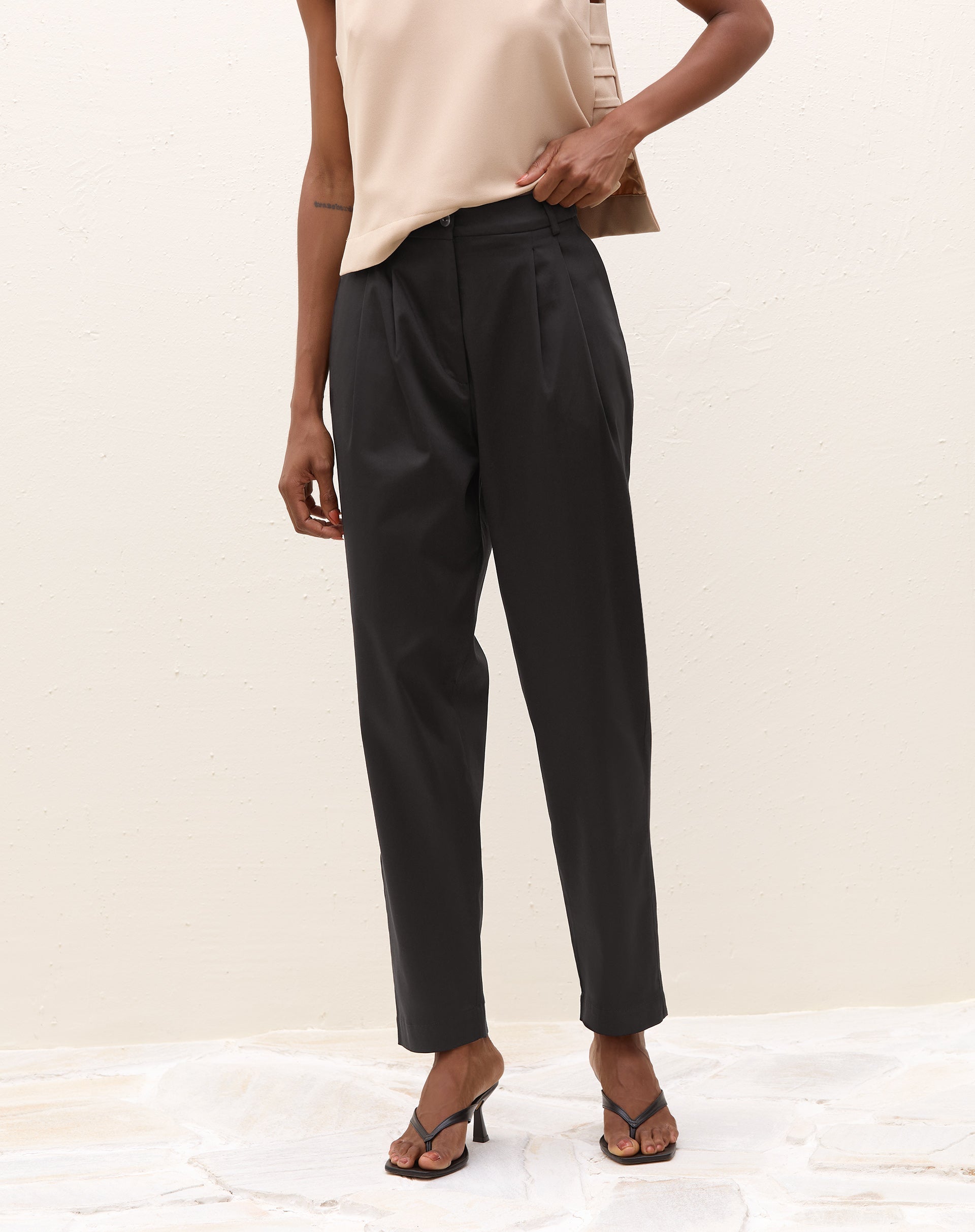 Modelo vestindo calça de alfaiataria baggy preta com pregas e blusa bege, vista lateral.