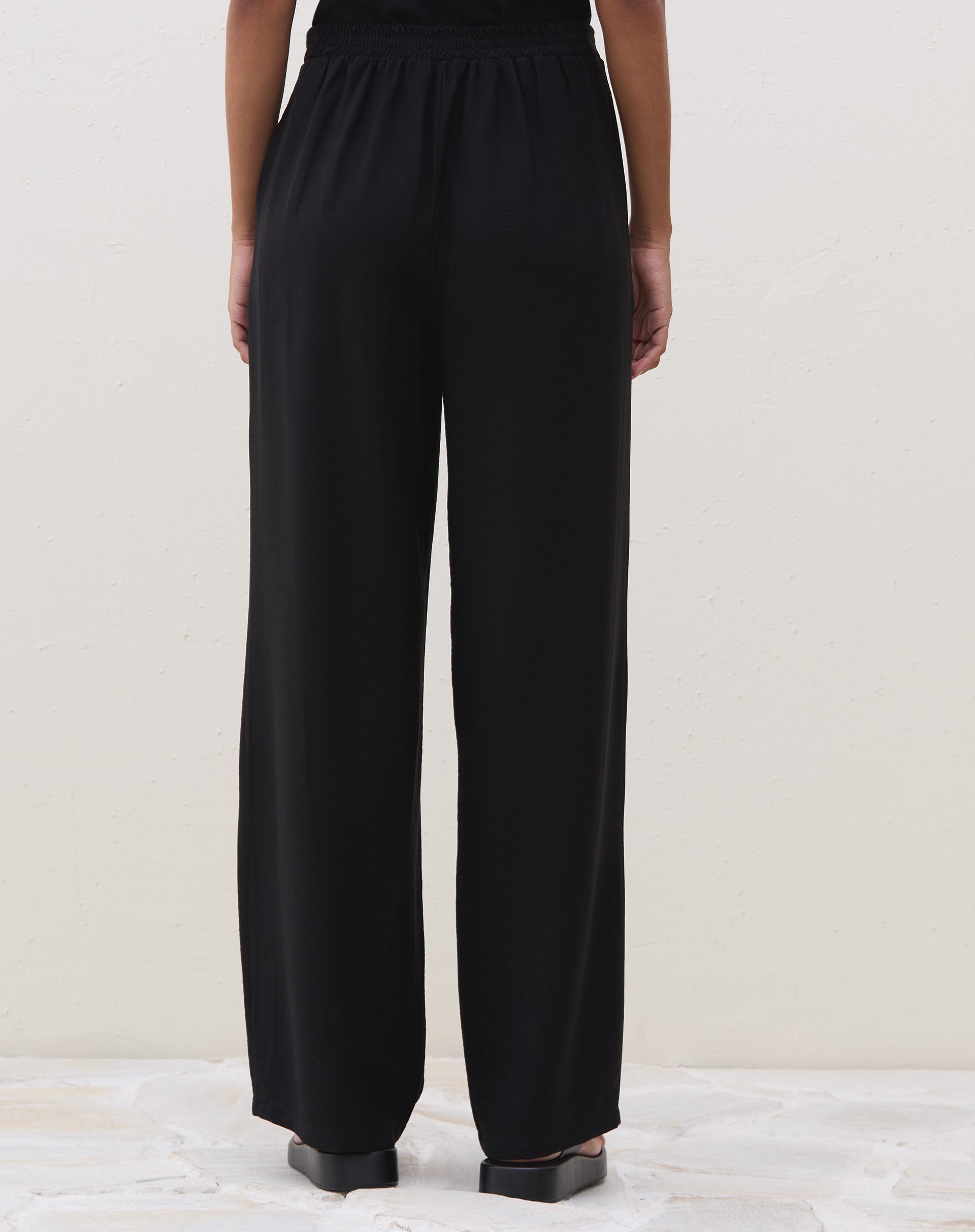 Modelo vestindo calça preta reta com cós elástico, combinada com blusa preta e sandália.