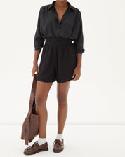 Modelo vestindo shorts preto com camisa preta e sapatos marrons, segurando uma bolsa.