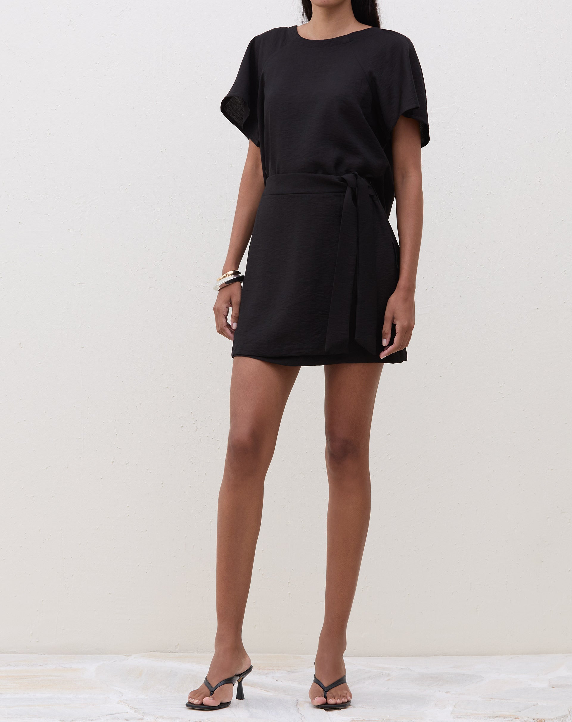 Modelo vestindo saia curta transpassada preta com amarração, vista frontal.