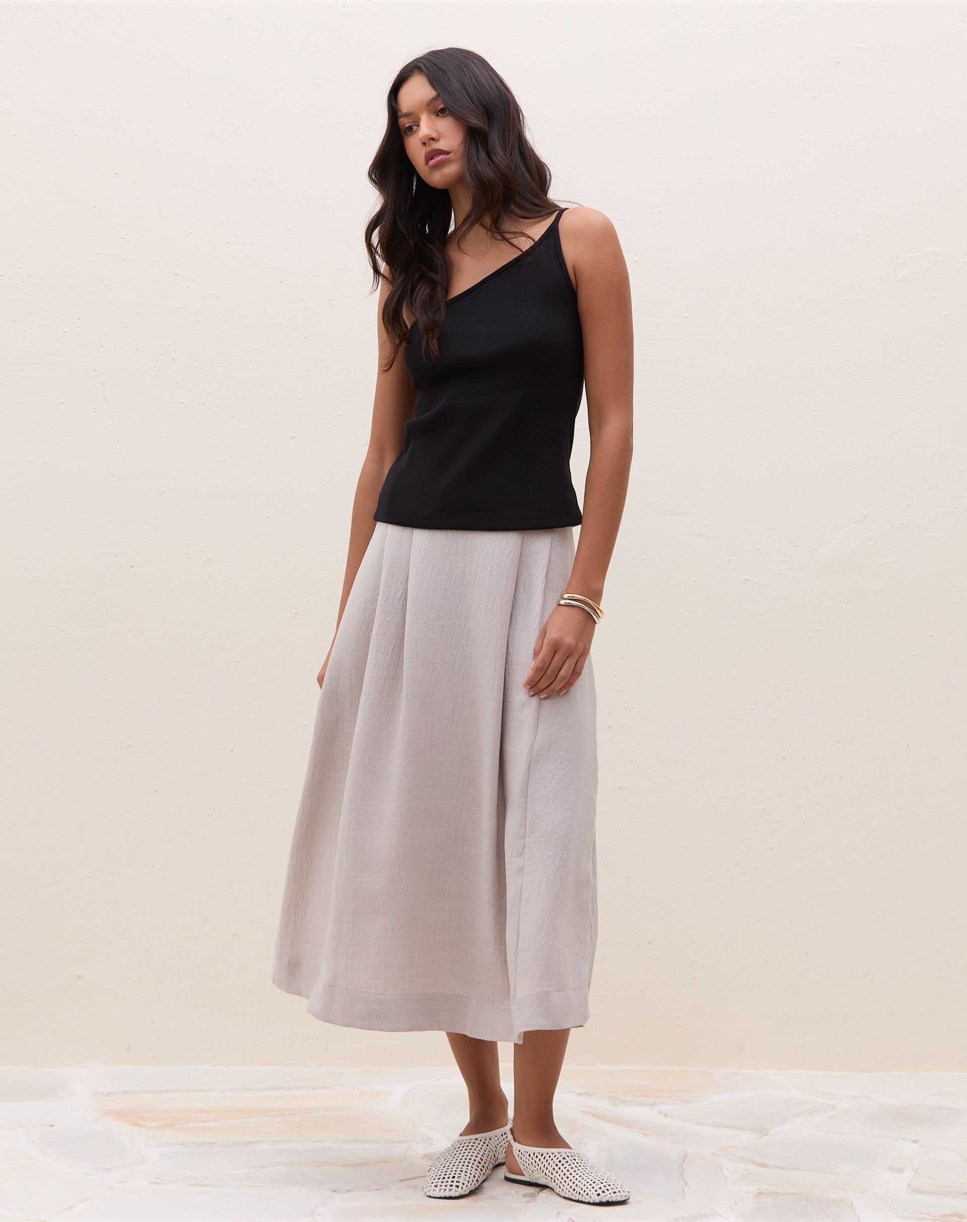 Modelo vestindo saia midi cinza claro com pregas, combinada com blusa preta e sapatos brancos.