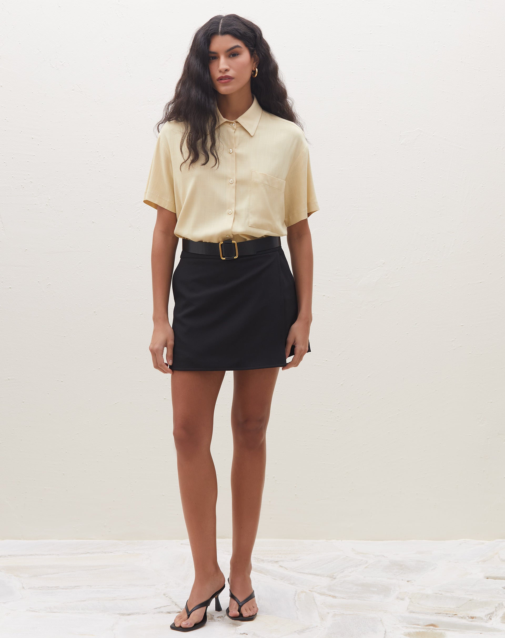 Modelo vestindo camisa manga curta com bolso em tom areia, combinada com saia preta e sandálias.