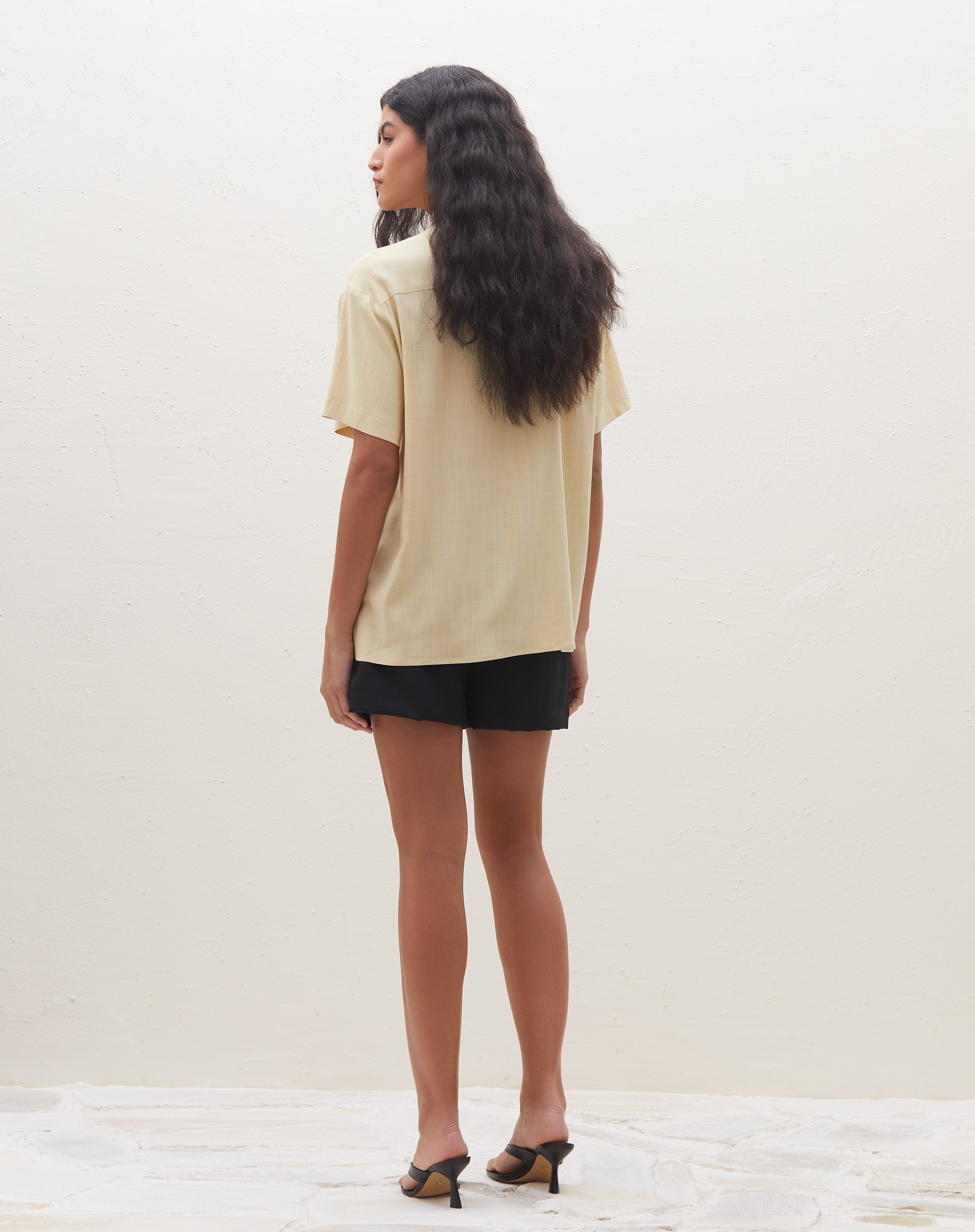 Modelo vestindo camisa manga curta com bolso em tom areia, vista traseira.