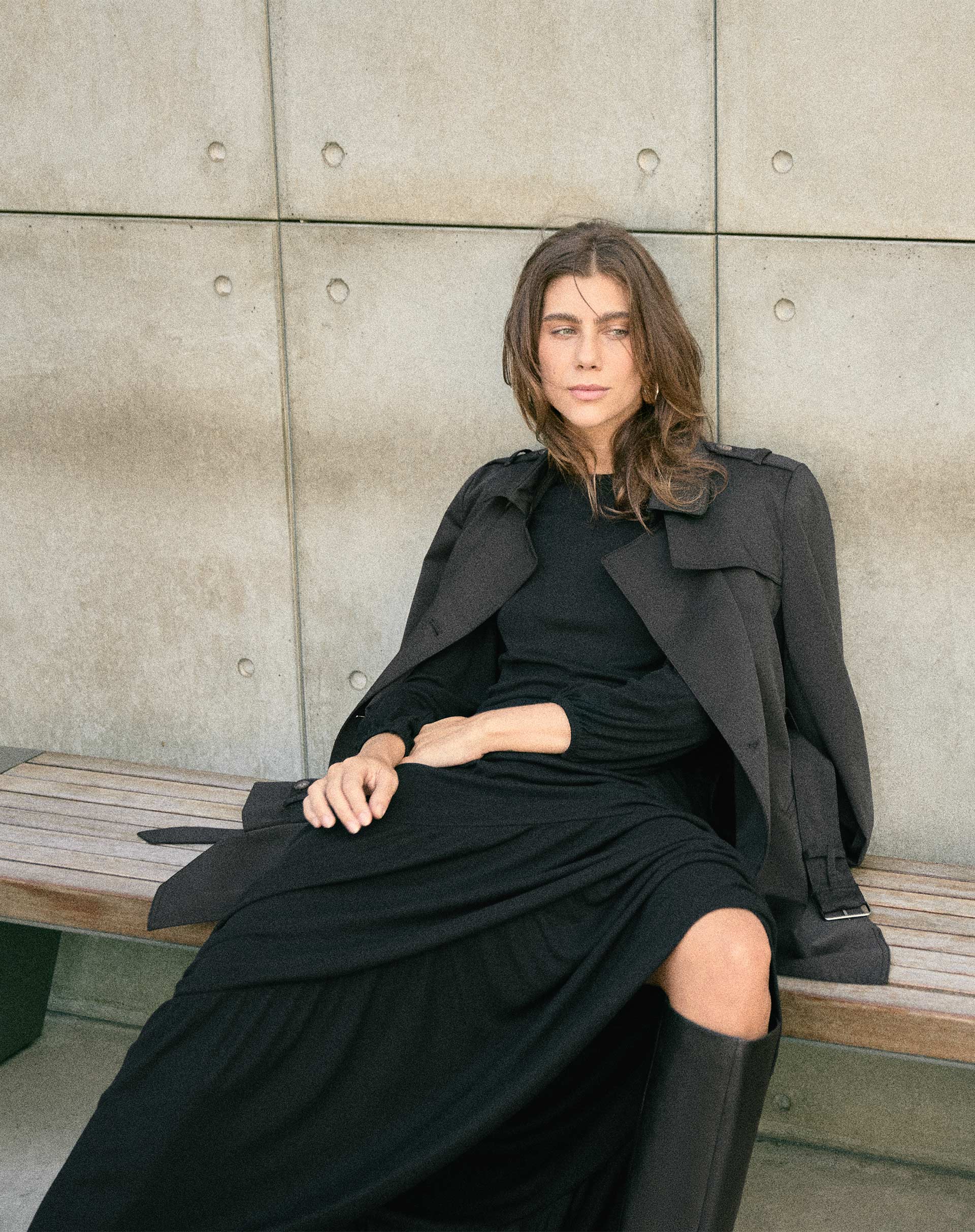 Modelo sentado em um banco vestindo um vestido longo preto com recortes de malha e um casaco preto.