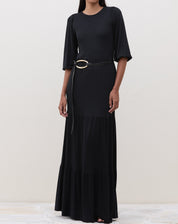 Modelo em pé vestindo um vestido longo preto com recortes de malha e cinto decorativo.