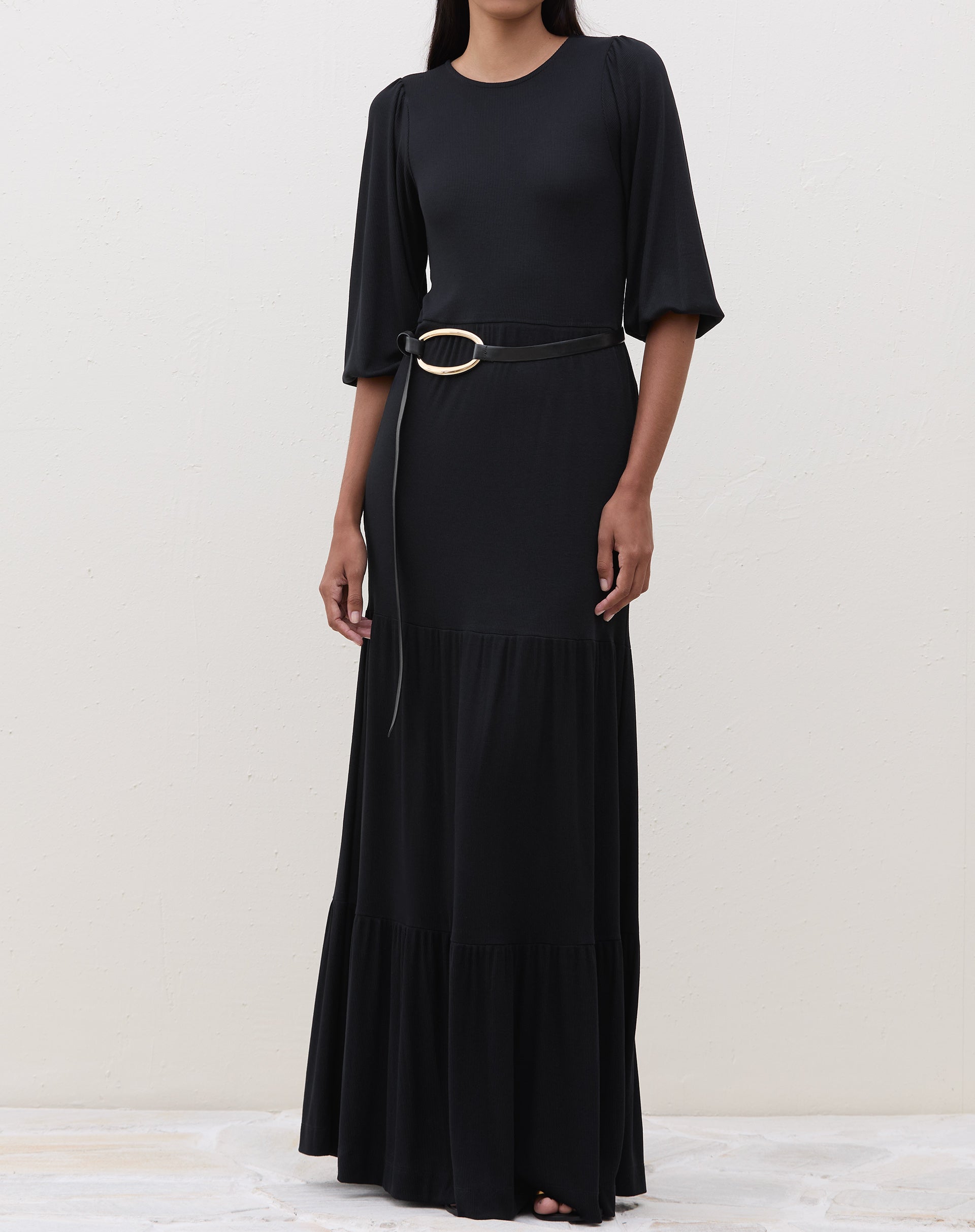 Modelo em pé vestindo um vestido longo preto com recortes de malha e cinto decorativo.