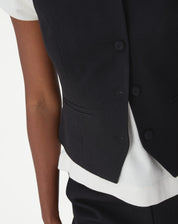 Close lateral do colete preto exibindo bolso paspa e recorte na barra com leve entalhe frontal.