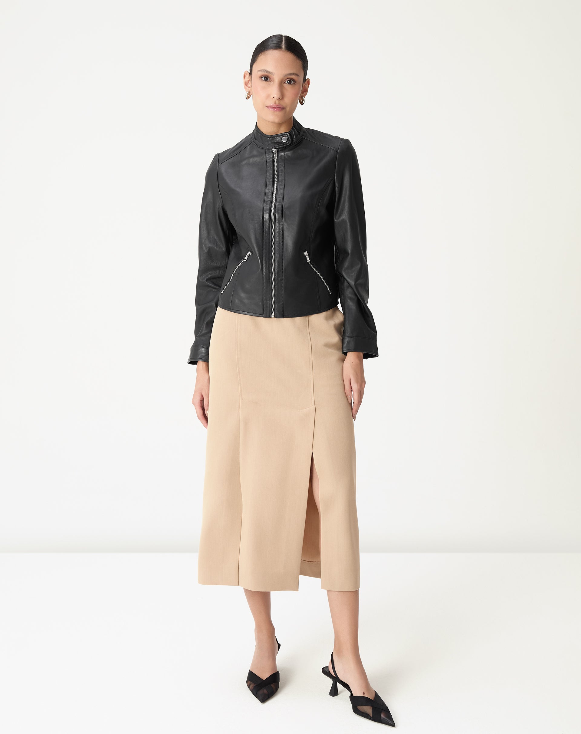 Modelo em pé vestindo saia midi alfaiataria bege com fenda frontal e cós médio, styling com jaqueta preta e sapatos pretos, vista frontal completa