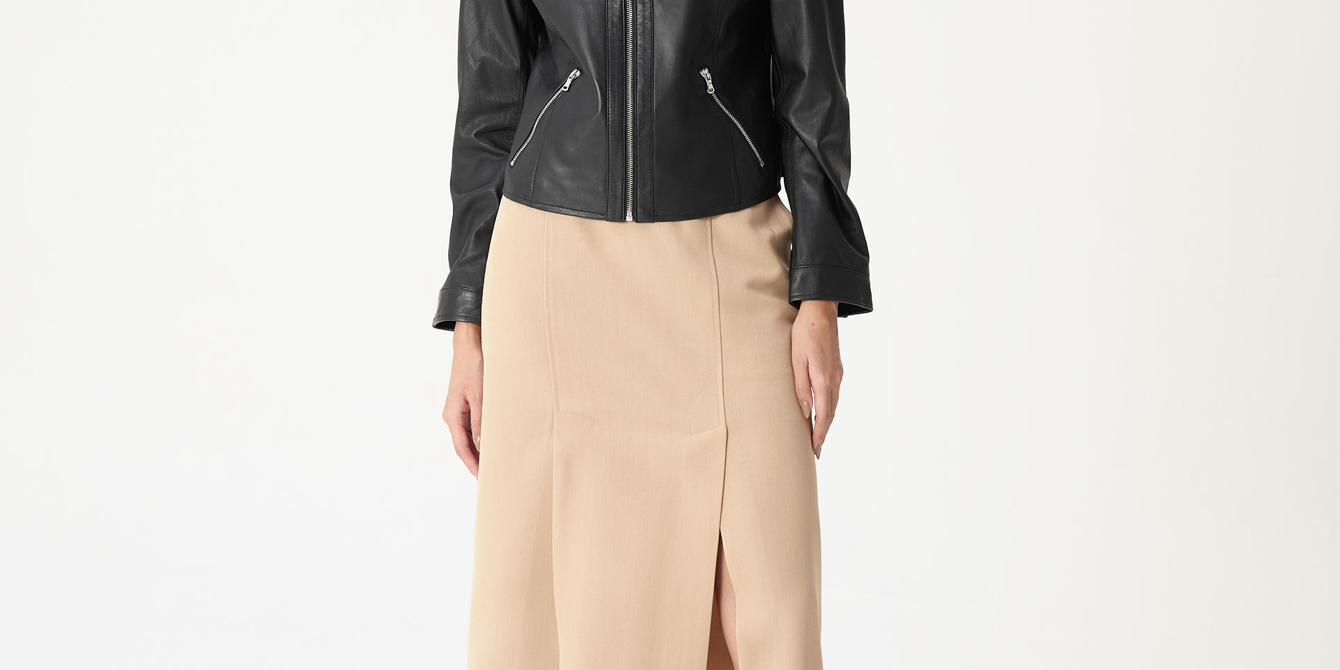 Modelo em pé vestindo saia midi alfaiataria bege com fenda frontal e cós médio, styling com jaqueta preta e sapatos pretos, vista frontal completa