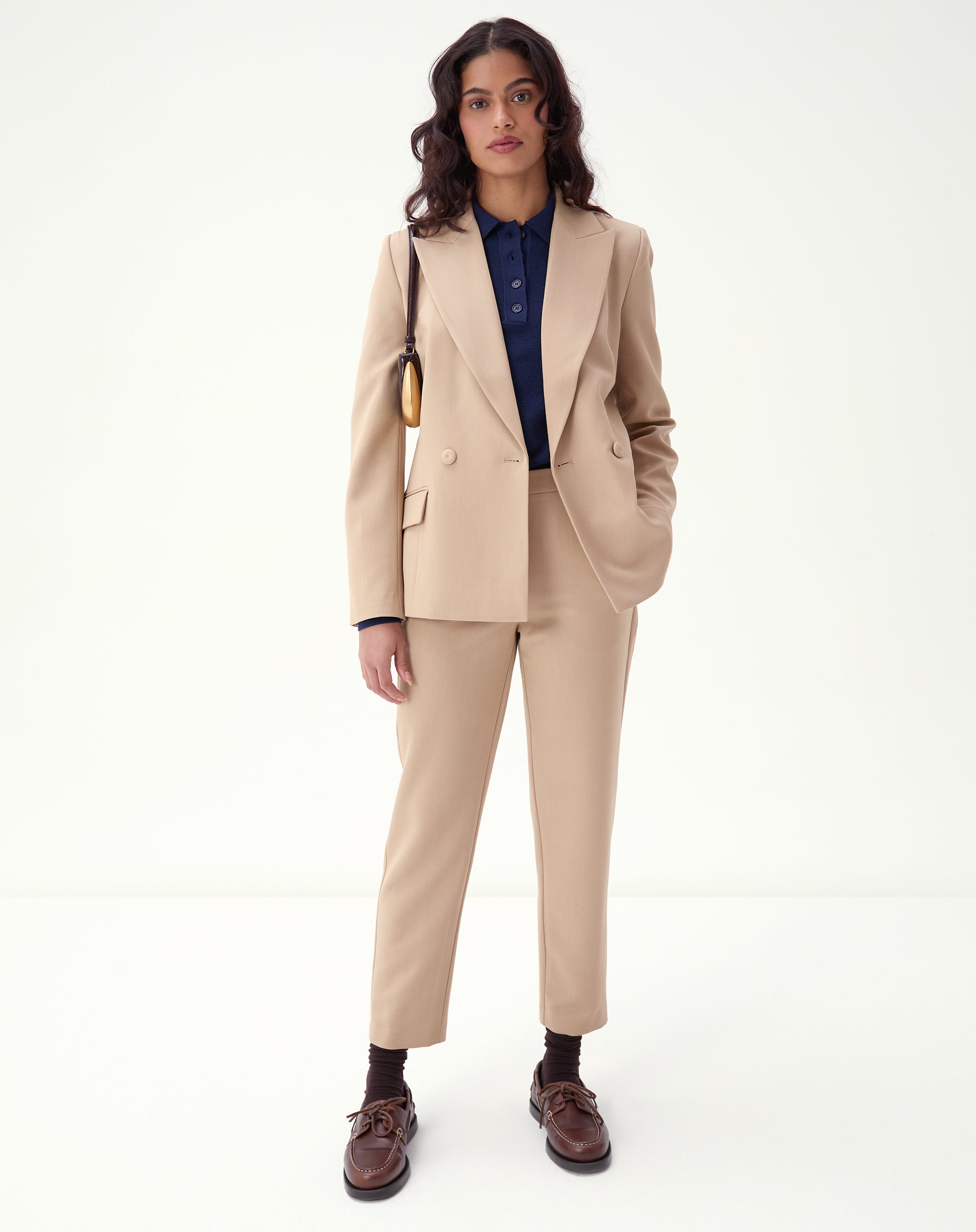 Mulher vestindo conjunto de alfaiataria bege com blazer e calça slim alfaiataria bege, look formal com blusa azul-marinho e sapatos marrons.